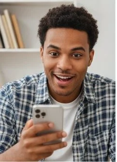 A surprised young Black man in a blue plaid shirt looks wide-eyed at his smartphone indoors, with books in the background.